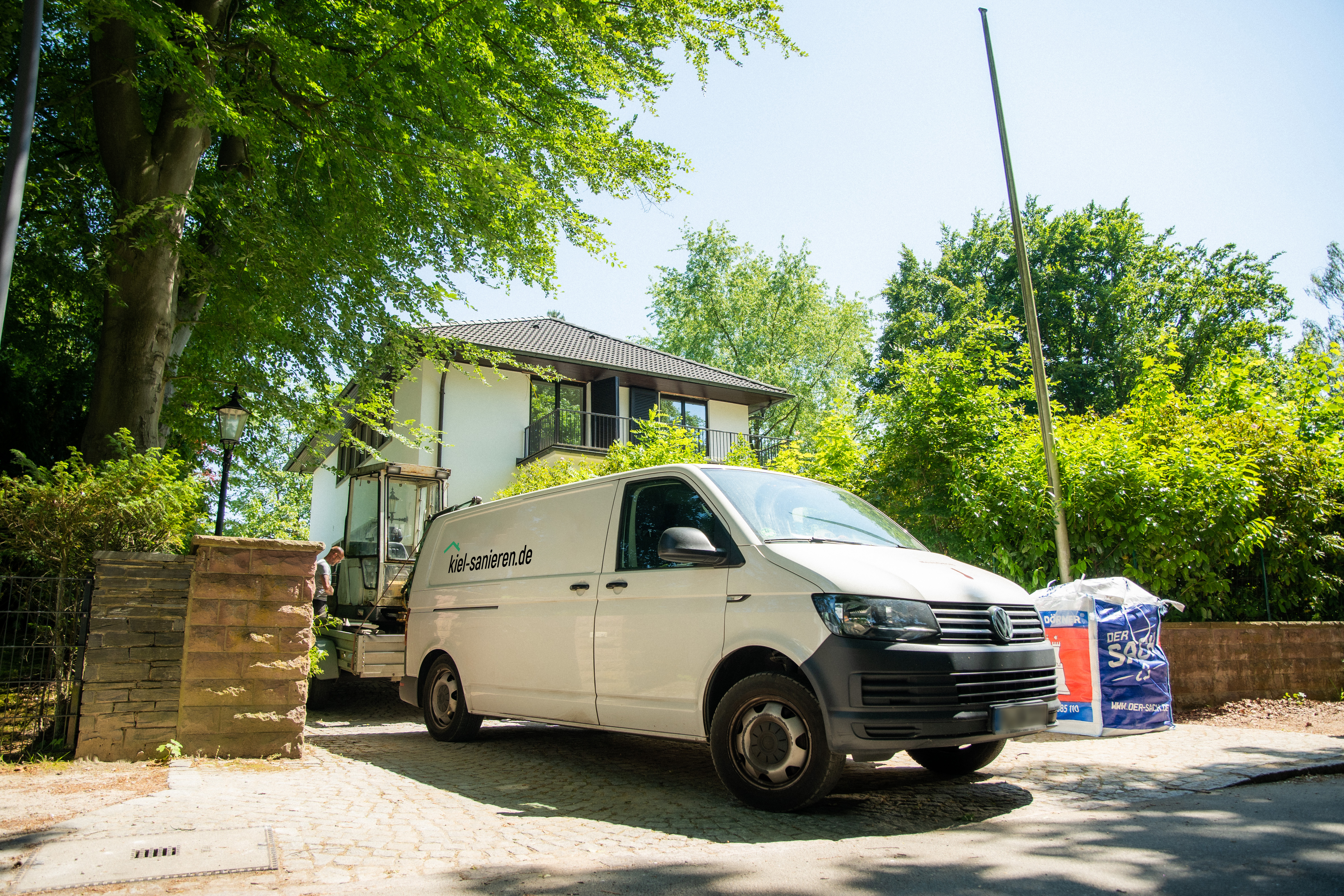 Transporter von kiel-sanieren.de vor einem Haus, bereit für Sanierungsarbeiten, umgeben von Bäumen und Baustellenmaterial.